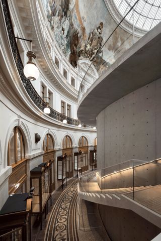 A detail of the hall at The Bourse de Commerce in Paris. We see many wooden doors to the left. To the right is a circular concrete structure that has white concrete stairs attached to it. We see renaissance art on the walls above.