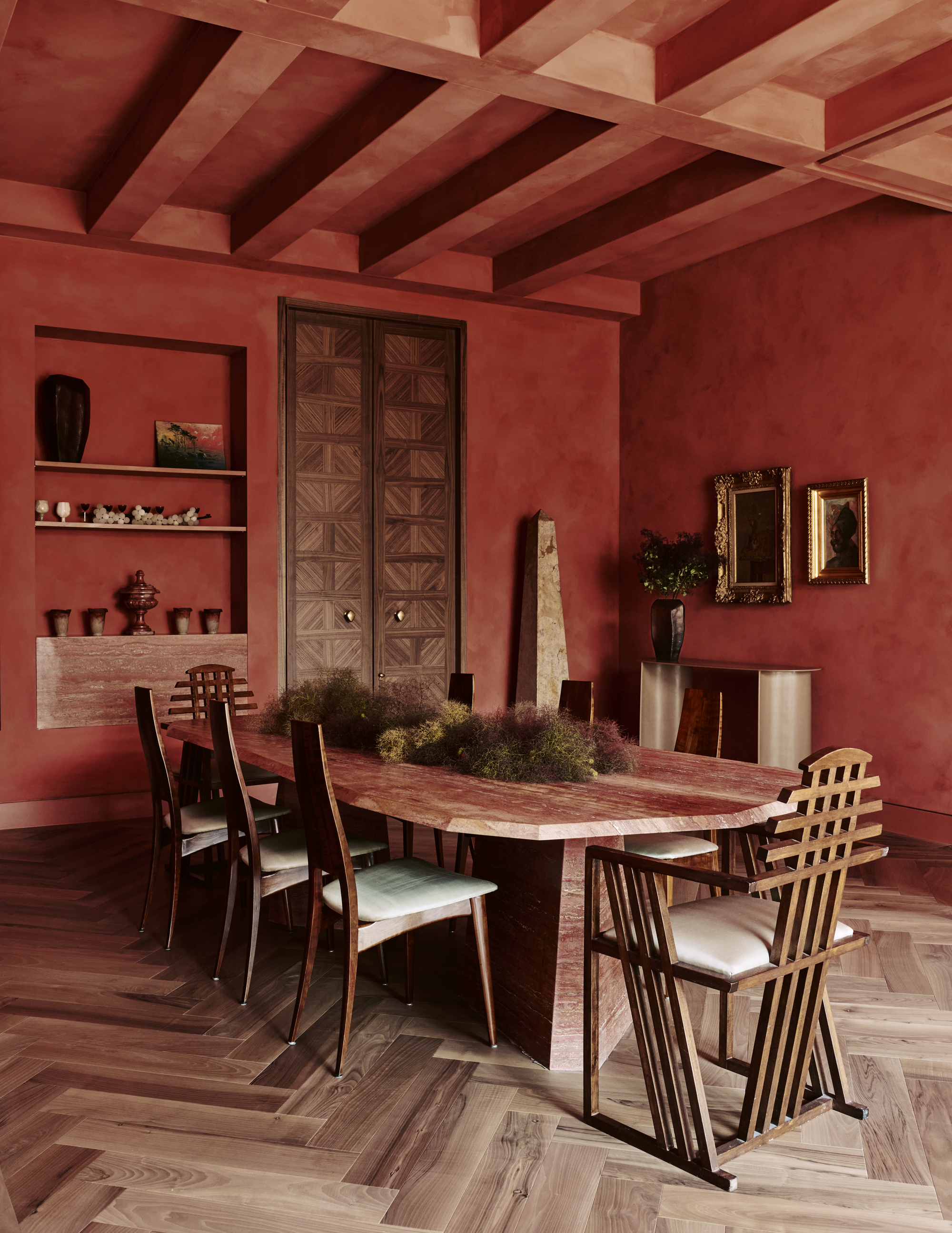 Image of a red, limewashed dining room with a marble table that has plants in the center and wooden dining chairs around it.