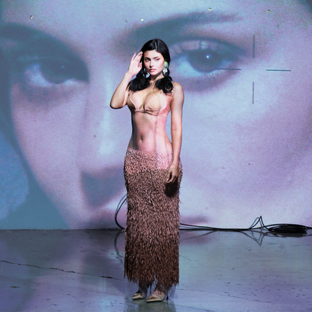 Kylie Jenner photographed by Aidan Zamiri for Who What Wear. She is wearing a Schiaparelli sheer look with nude bra and textured skirt. She is standing in front of a projected image of herself. 