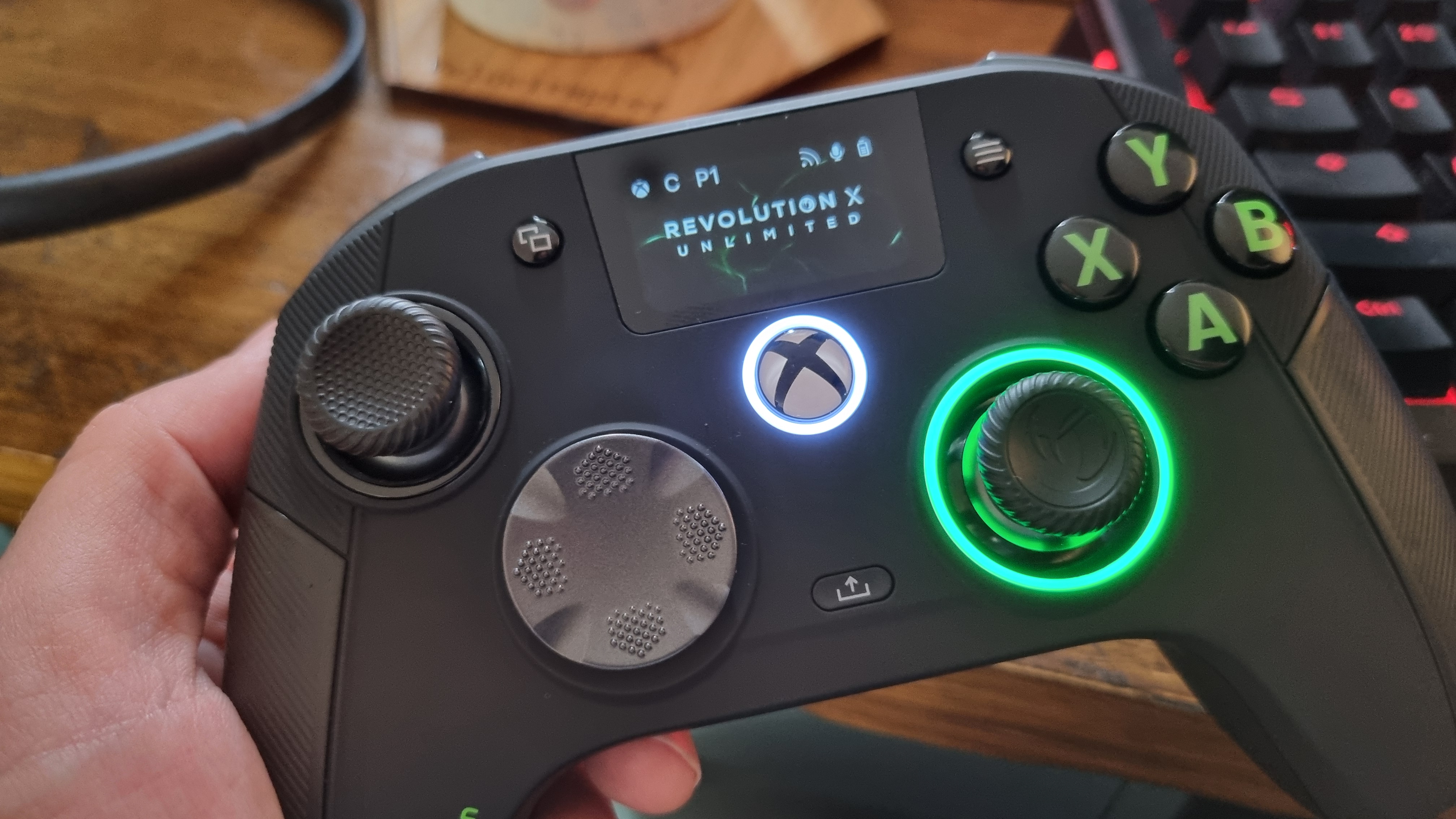 The Nacon Revolution X Unlimited controller with its thumbstick ring lit up in green, held in the hand in front of a wooden desk