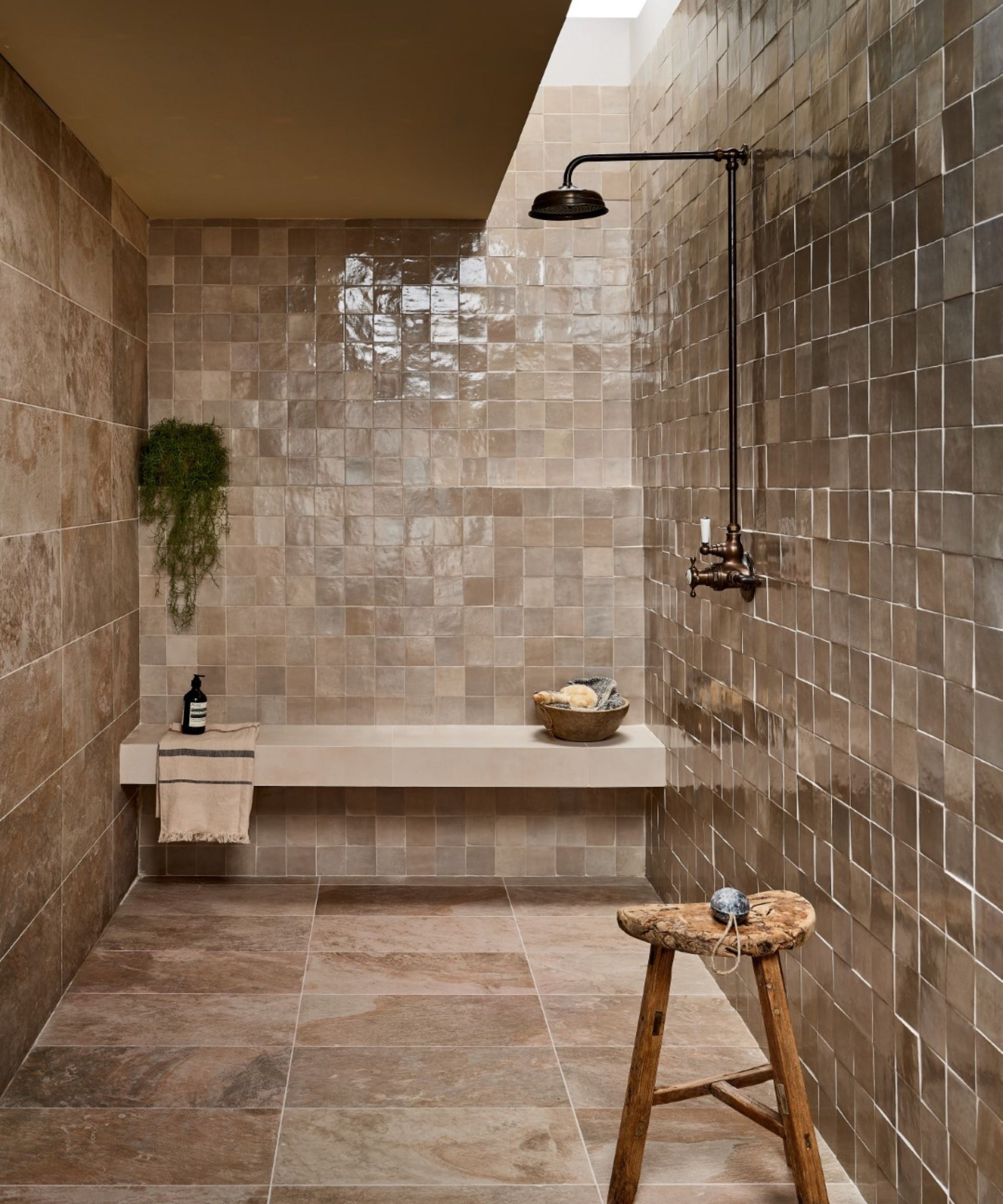 A spa-like open shower room entirely clad in warm taupe zellige square tiles on the walls and large-format stone-effect floor tiles. A bronze wall-mounted rain shower head, floating plaster bench, hanging trailing plant and rustic wooden stool create a serene, earthy atmosphere lit by a ceiling rooflight.