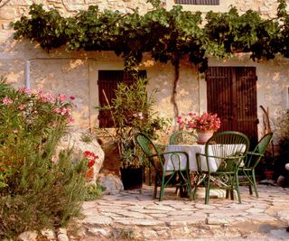 Green chairs and table on a stone terrace in front of a rustic stone cottage with climbing plant