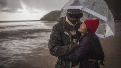 A couple huddle under an umbrella together on a cold, rainy beach.