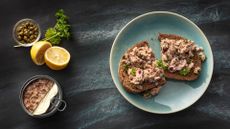 A slice of bread topped with tuna salad sits on a blue plate on a dark surface. An open can of tuna, fresh herbs and lemons sits to the left.