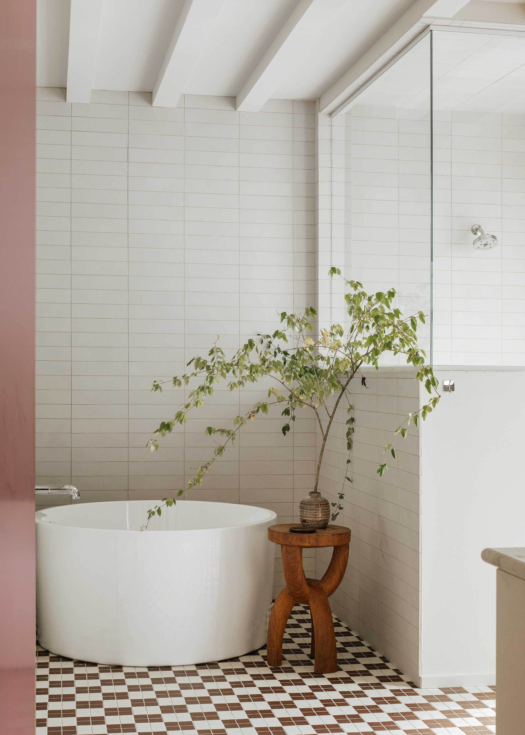 A white bathroom with horizontal metro wall tiles, checkerboard floor tiles, a round tub bath and a sculptural wood side table topped with a branch in a small vase.