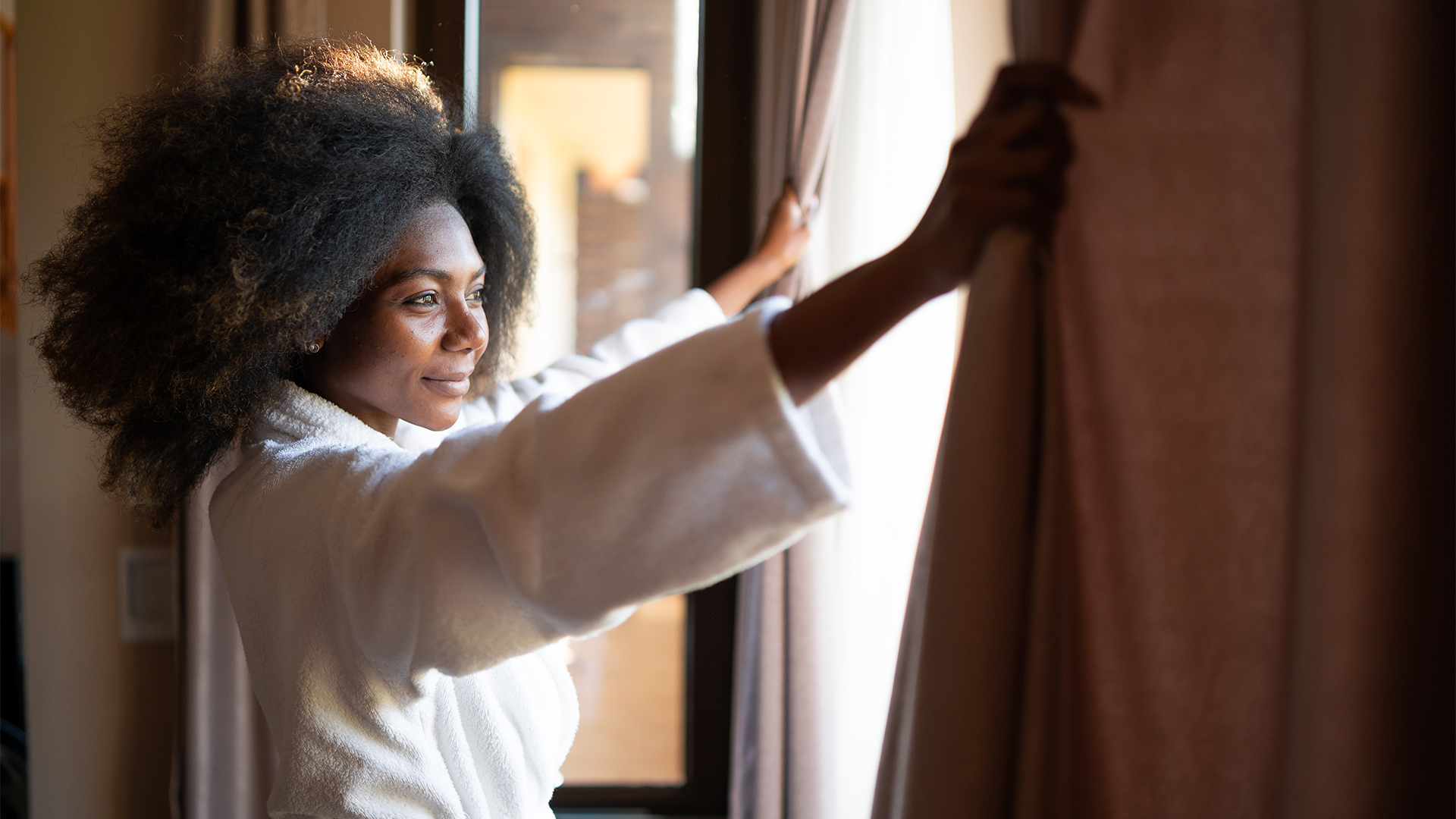 A woman stands in front of a window in her bedroom during time, with her hands on the curtains preparing to pull them closed