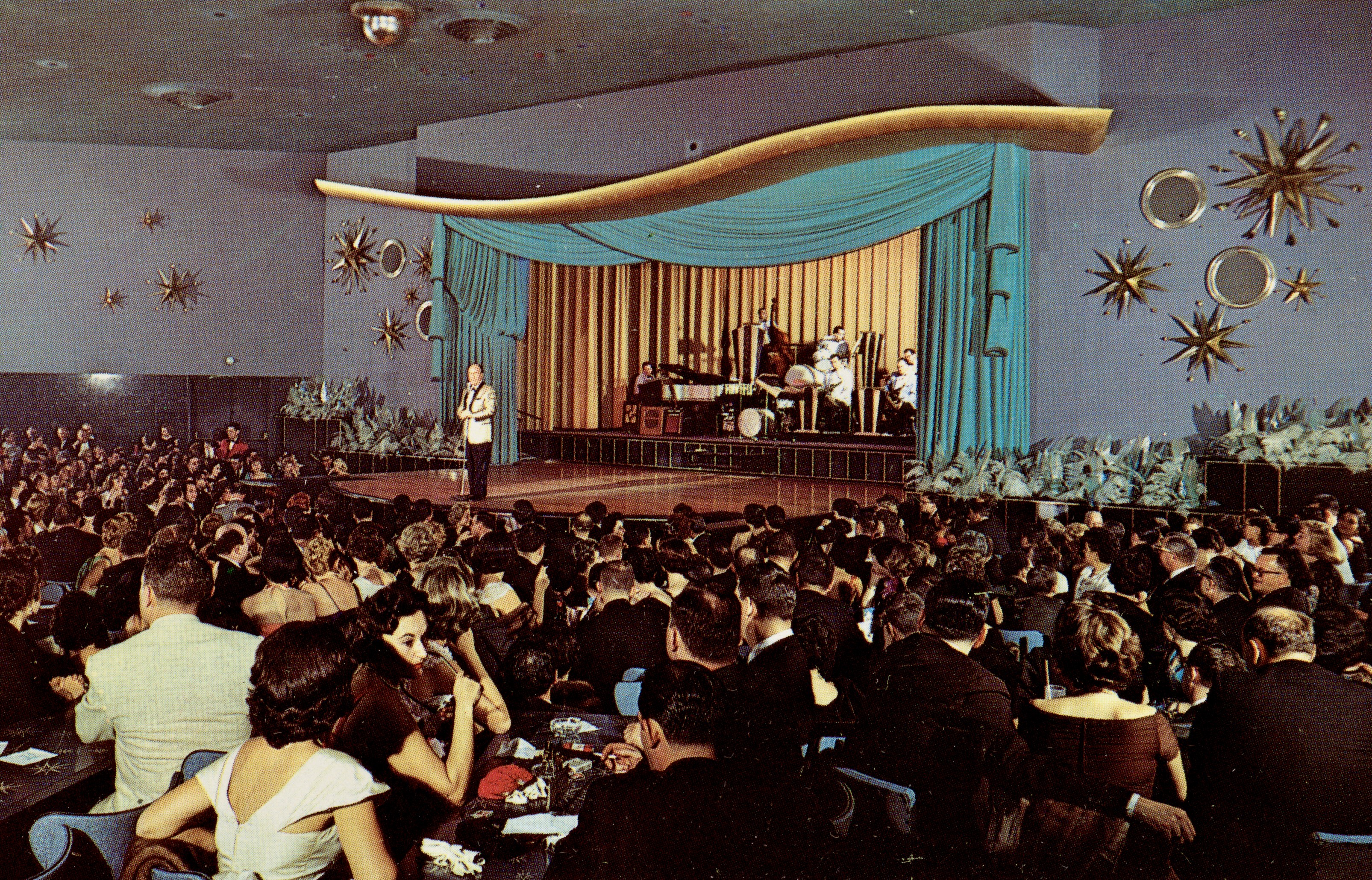 A 1950s photograph of a mid-century modern theater situated in a hotel portrays a crowd sitting in the audience as a performance goes on in the background. Decorating the walls are golden stars and circles, which surround an ocean green curtain.