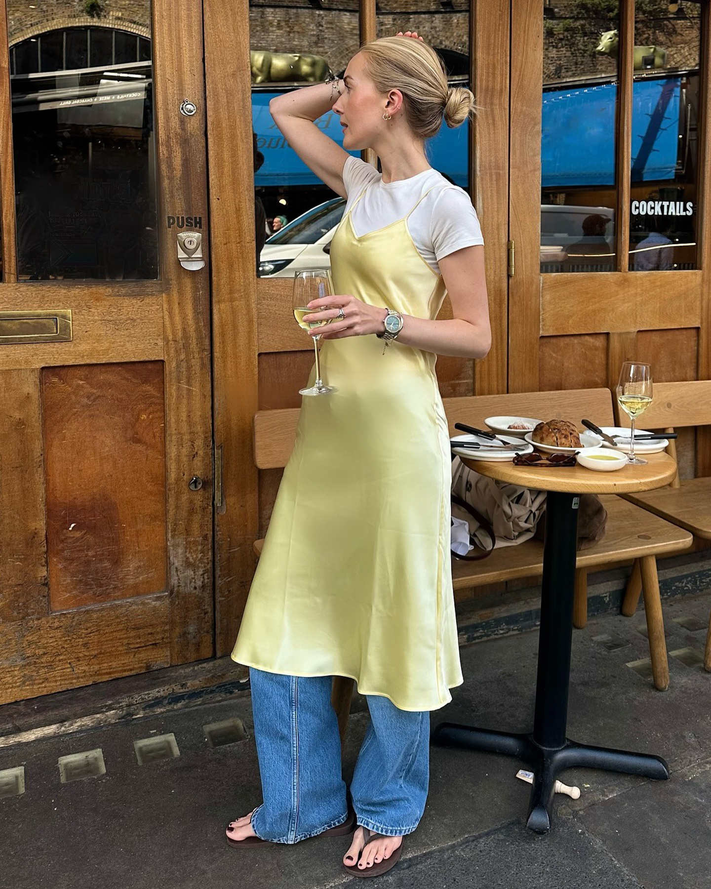 British influencer Chloe Butler poses with a glass of white wine outside of a London restaurant wearing a yellow slip dress over a white t-shirt, jeans, and brown flips-flop sandals.