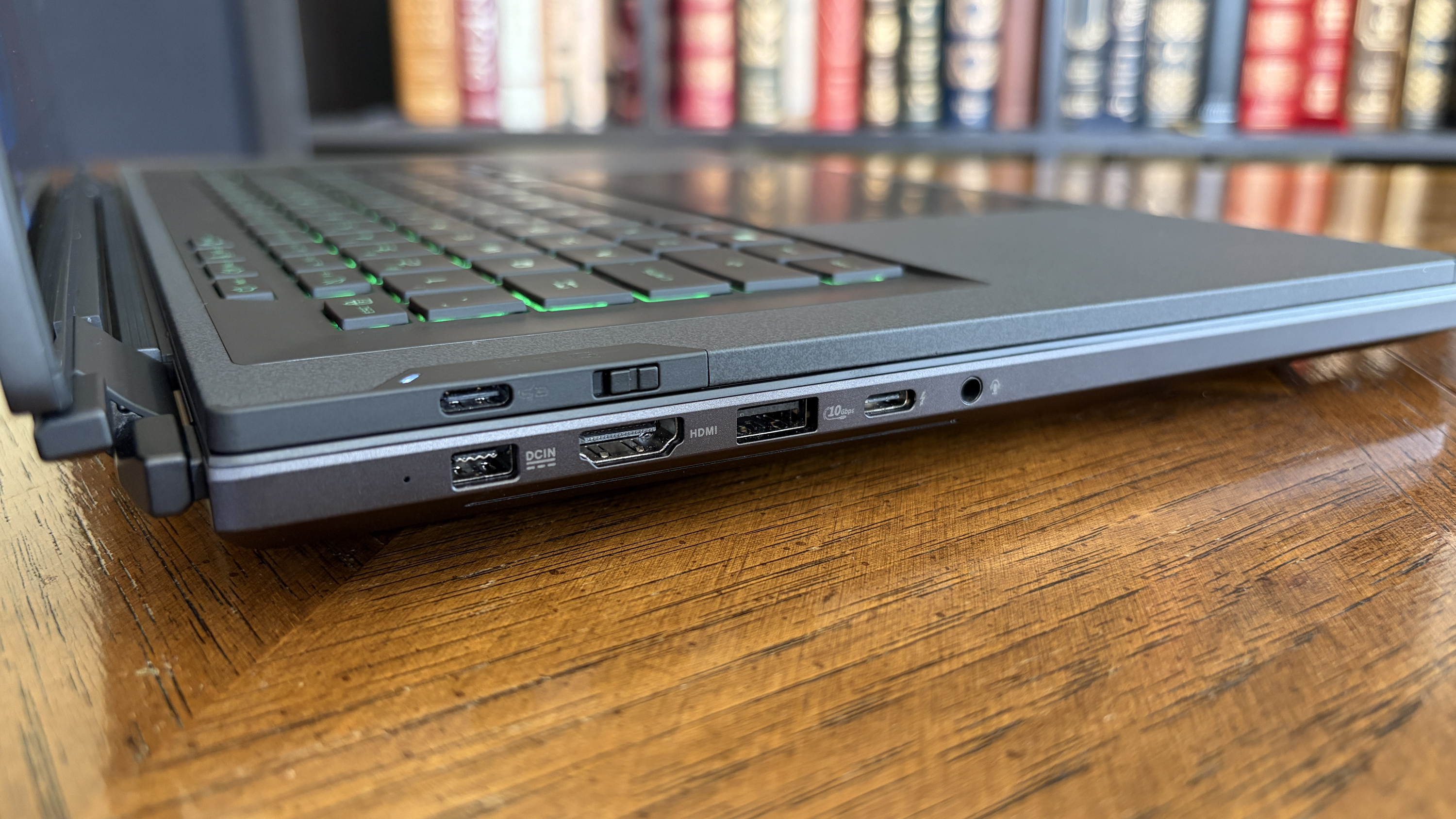 A close-up of a laptop's side view on a wooden table, showing multiple ports including USB, HDMI, and headphone jack, with books blurred in the background.