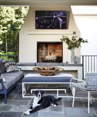 outdoor living area surrounded around a large outdoor working fireplace and wall mounted TV, with a tiled patio area with sofa, ottoman and chairs plus a black and white dog