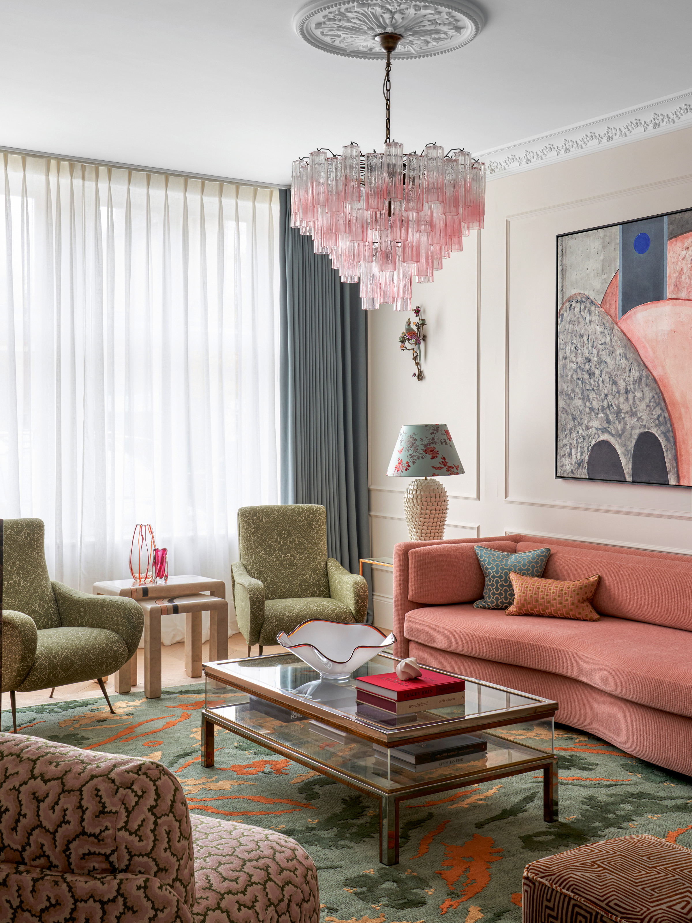 Living room with white walls, coral sofa, green armchairs, green and coral rug, glass coffee table and pink chandelier