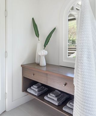 Relaxed, minimalist style bathroom with dark wood shelving unit, open and closed storage, low open shelves storing white towels, two decorative vases with leaf stems, arch window, white painted walls.