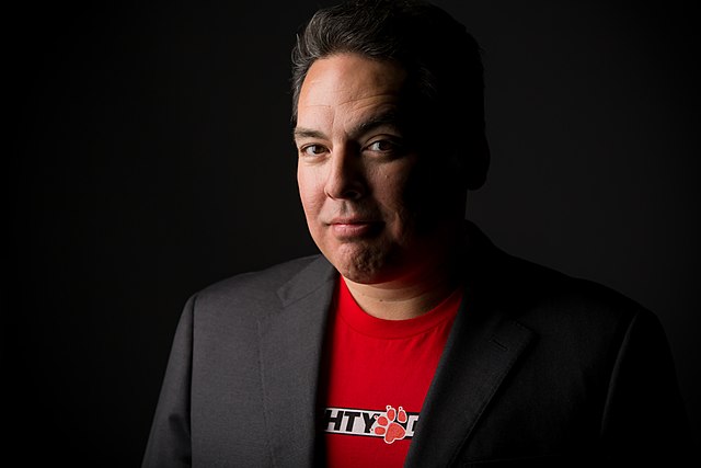 Shawn Layden stands in dramatic lighting against a black background, wearing a dark suit and red &ldquo;Naughty Dog&rdquo; T-shirt, conveying confidence and focus.