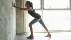 A woman stands in an empty room with a large window stretching against a wall. Her arms are straight, hands on the wall, and she is in a staggered stance so her right leg is behind her and her left leg slightly forward. Her torso leans forward as she bends into the stretch.