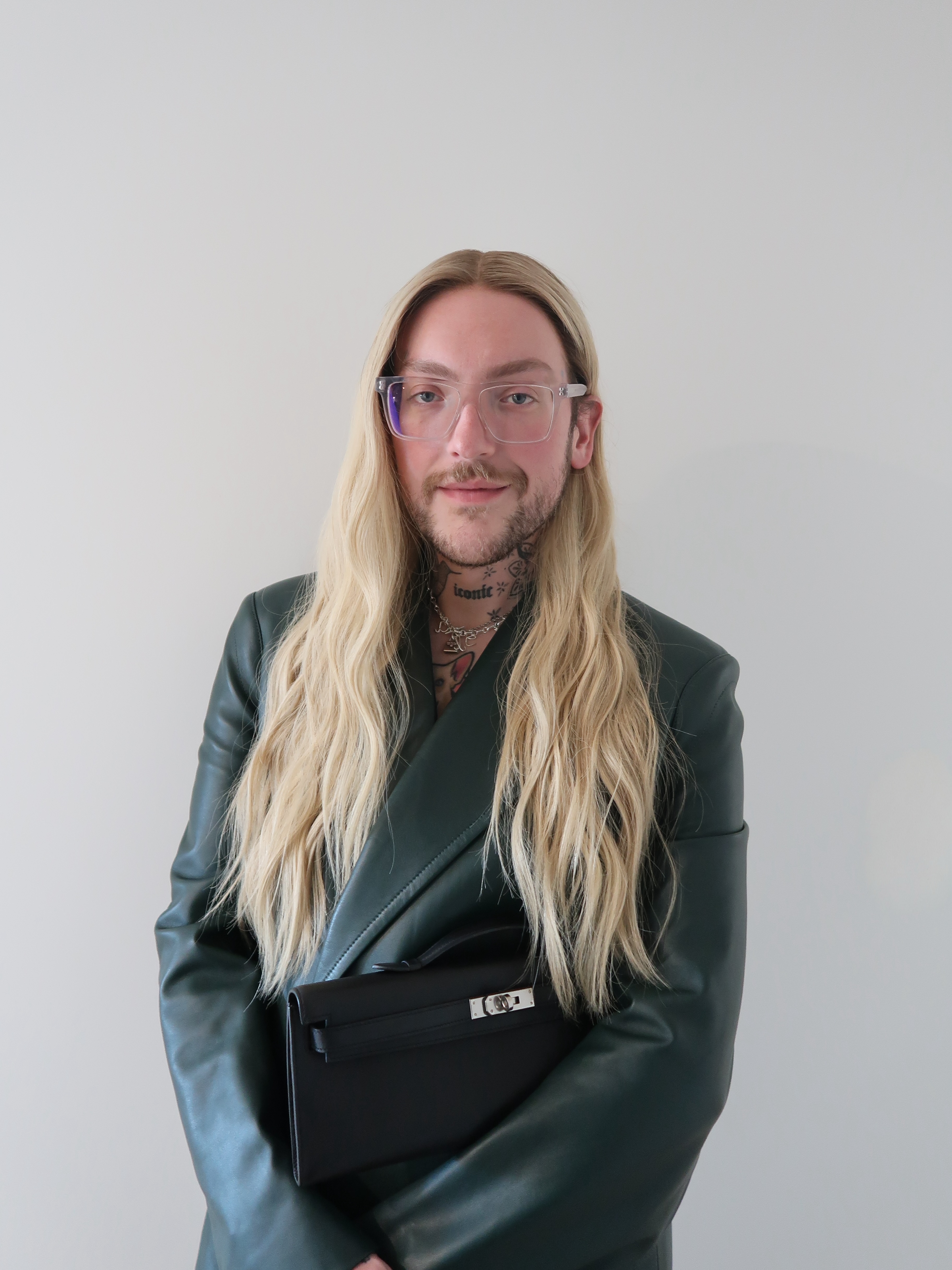 a headshot of a white man with long, blonde hair wearing a dark, leather blazer in front of a white backdrop