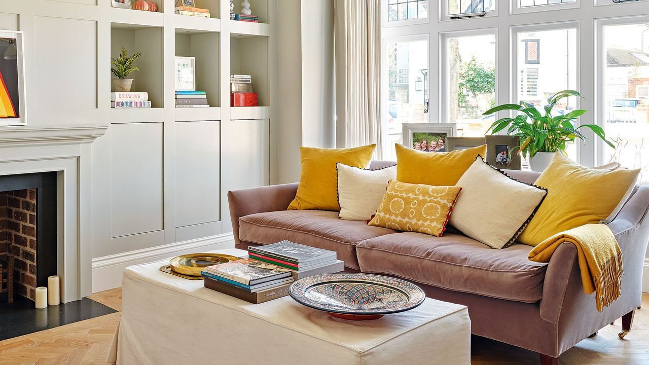 A white living room with a wall of windows, built in storage and a muted pink sofa.