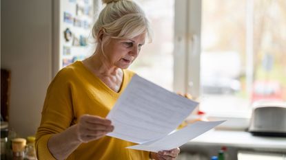 An older woman looks at some paperwork while standing in her home.