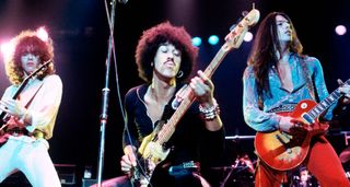 Thin Lizzy live in 1978, Brian Robertson in white, playing his Les Paul, Phil Lynott in the middle on bass and vocals, and Scott Gorham in red trousers and an open blue shirt playing a tomato burst Les Paul