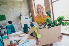 Photo of a sad professional woman who has been laid off. She is holding a box of her office supplies and plants as she leaves her desk.