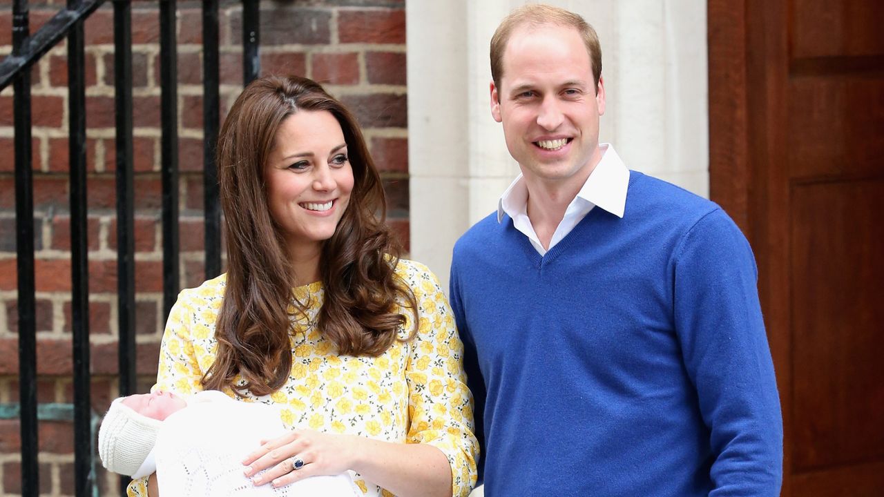 Princess Kate stands outside the hospital holding Princess Charlotte who is wrapped in a white blanket, while Kate wears a yellow floral dress and Prince William wears a blue sweater