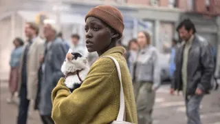 Lupita Nyong'o holds a cat and wears a yellow coat walking a street covered in rubble in 'A Quiet Place: Day One'