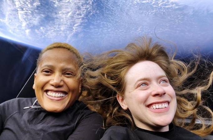 two women floating on a spacex spacecraft with a window in behind showing a view of earth