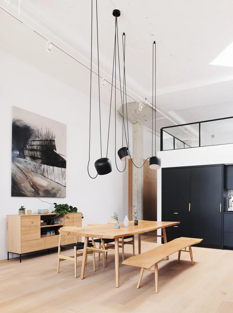 SoMa Loft by Malcolm Davis Architecture (MDa) features a wood table with wood chairs, a wood side board with crockery, a painting on the wall and black kitchen cupboards.