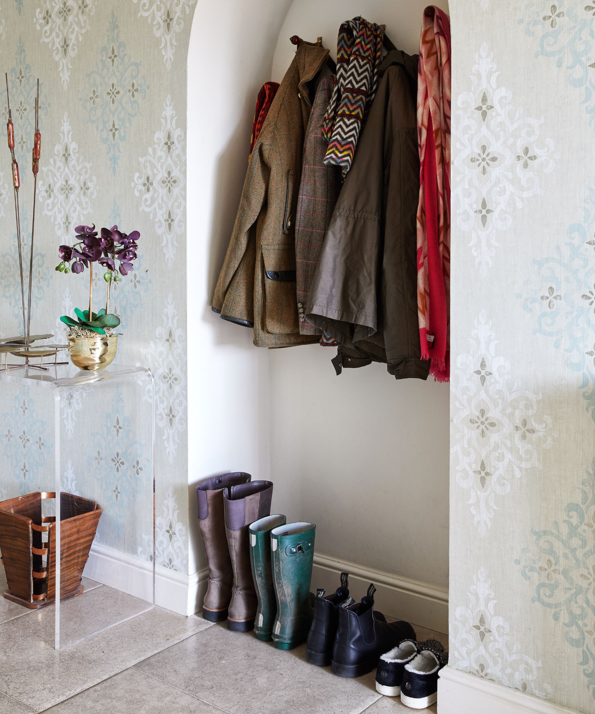 A row of shoes lined up in a wallpapered mudroom underneath a coat rack in a white alcove. To the left is a clear acrylic table with a flower in a gold plant pot on top, and a brown bin beneath.