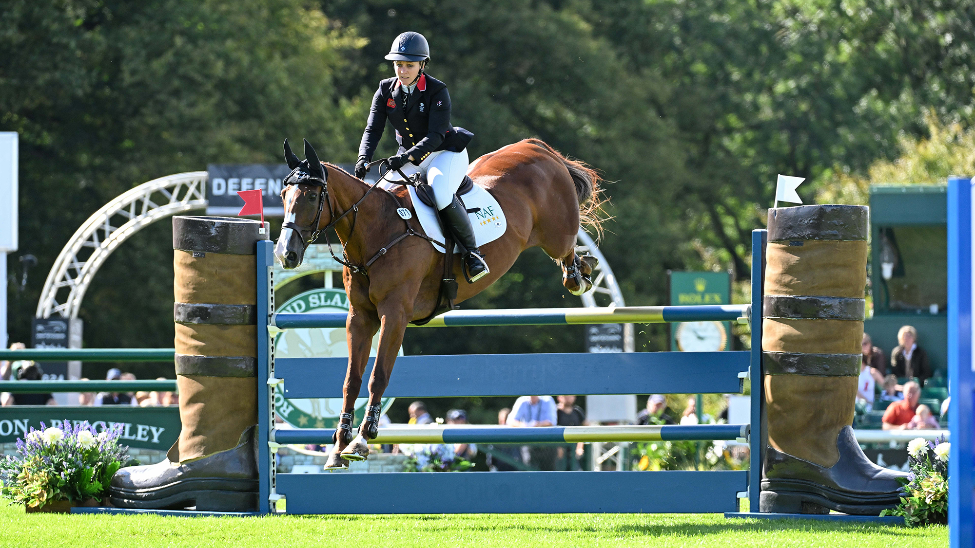 Gemma Tattersall, a rider in a black coat, pilots chestnut horse Chilli Knight over a fence with wings shaped like boots at Burghley 2024