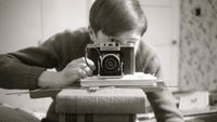 Black-and-white self-portrait with an Agifold camera, on top of a stack of books obscuring the photographer's face, pointing at a mirror 