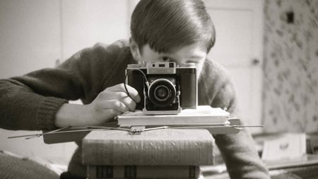 Black-and-white self-portrait with an Agifold camera, on top of a stack of books obscuring the photographer's face, pointing at a mirror 