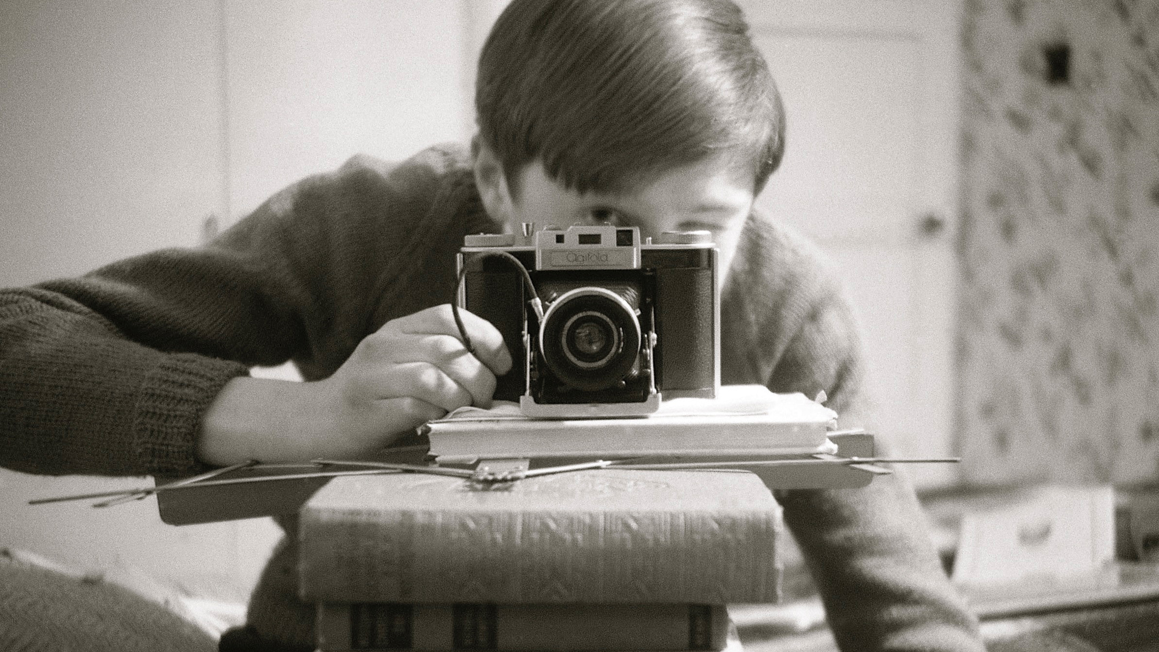 Black-and-white self-portrait with an Agifold camera, on top of a stack of books obscuring the photographer's face, pointing at a mirror  