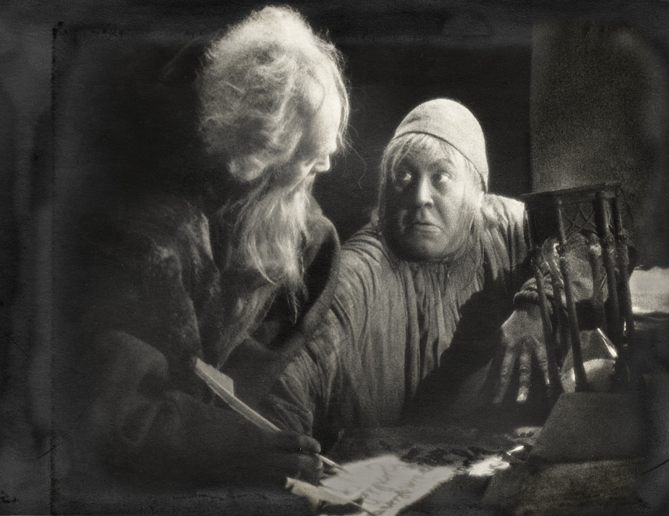 An old man with a long white beard and wild hair signs a parchment with a quill, while a wide-eyed figure in a skullcap leans in beside him, one clawed hand resting on the table next to an hourglass.