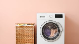 A washing machine with clothes inside next to a full wicker laundry hamper on a pink background