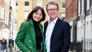  Davina McCall and Nicky Campbell standing in a city street, Davina wears a black roll neck with a green jacket, while Nicky has a white shirt open at the neck and a navy blazer on.