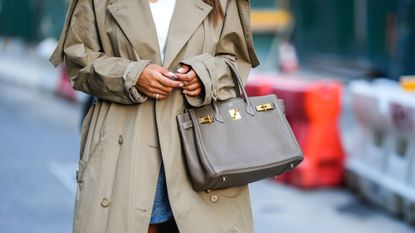 Tamara Kalinic wears quiet luxury nails, white wavy neck top, a beige long trench coat, a blue denim short skirt, a gray taupe Birkin handbag from Hermes