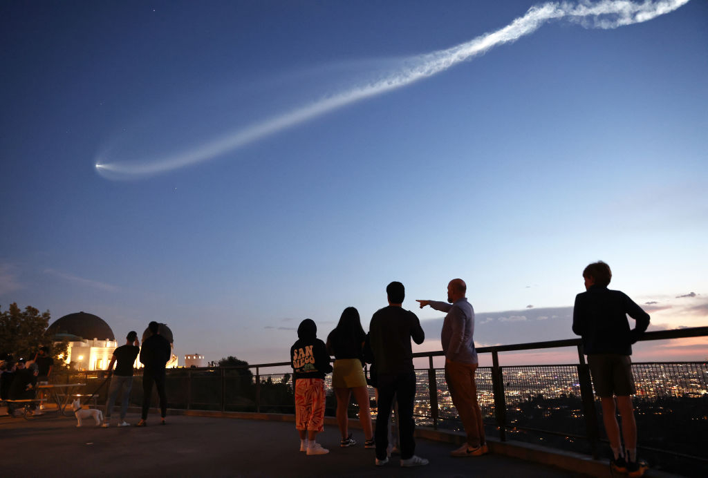 an expanding white plume extends from a small point of white light on the left, growing thicker in a stream across the darkening blue sky. Silhouettes of onlookers line the bottom, near a guard rail fence. City lights are seen low in the distance.