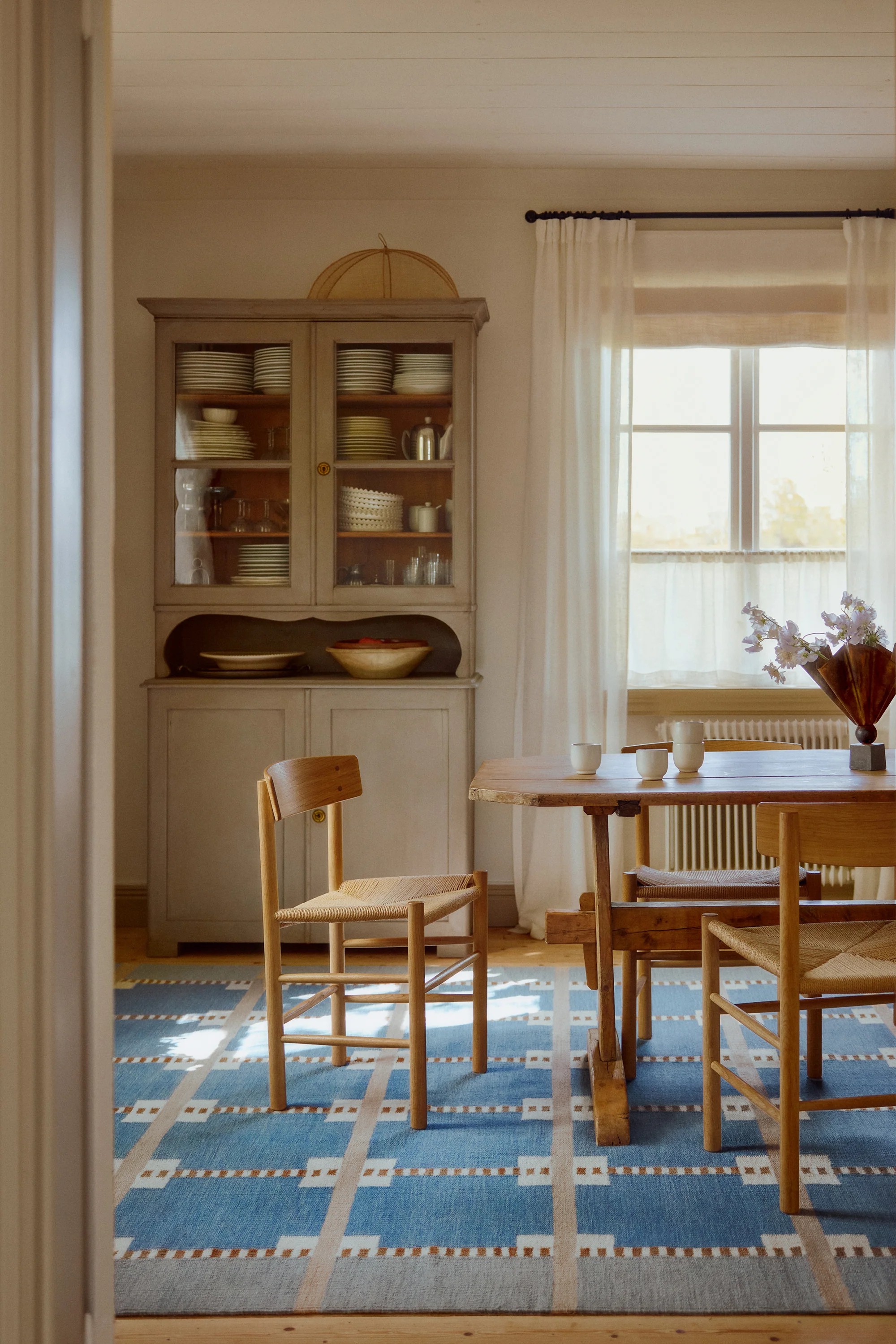 Image of a modern rustic dining room with a wooden table and chair set, styled on top of a blue, geometric area rug. There is a window with white curtains on the back wall. Beside the window is a storage cupboard with dishes inside.