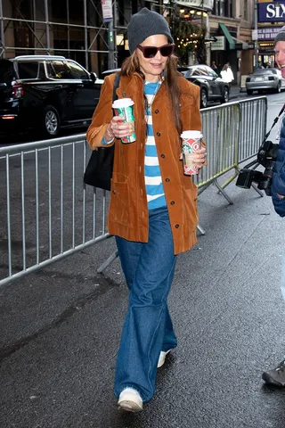 Katie Holmes is wearing a gray beanie, blue striped shirt, tan suede jacket, denim jeans, and white sneakers.