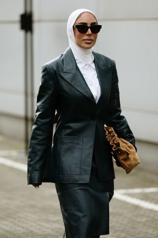 A woman at Paris Fashion Week wearing a black leather blazer, white shirt, black skirt, white head scarf, and brown bag