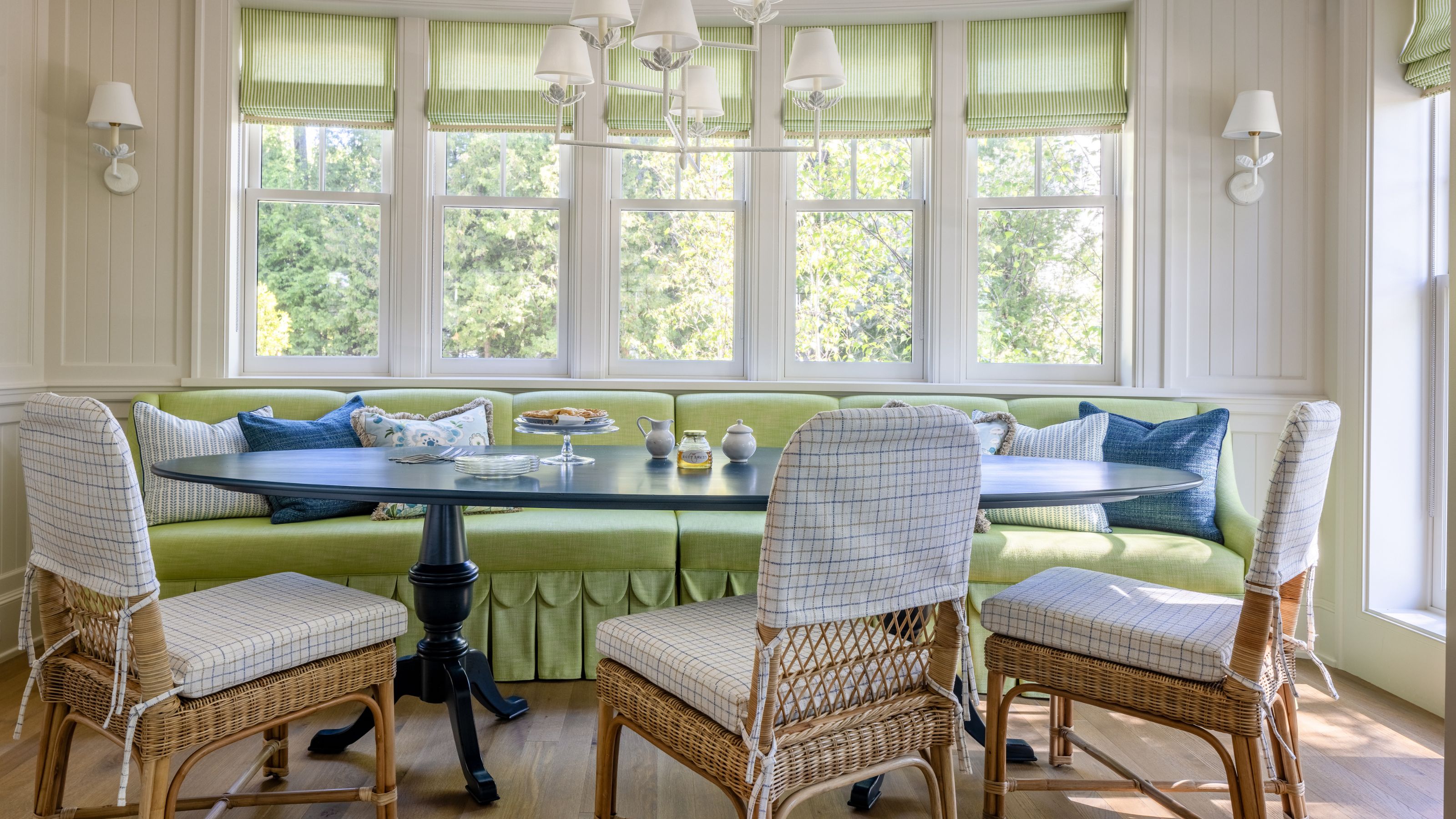 a neutral breakfast area with a green scalloped skirted banquette, large windows, wooden table and rattan and plaid chairs