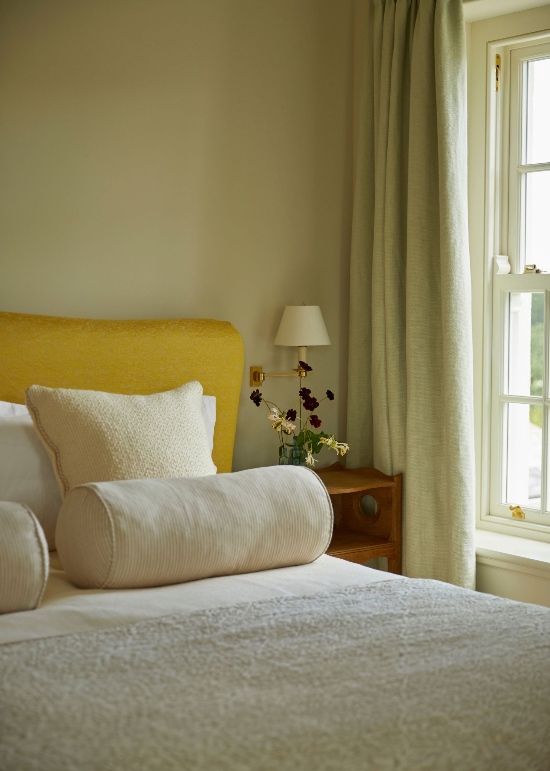a neutral bedroom with soft butter-yellow walls, a lemon-yellow headboard, layered ivory bedding, and light neutral curtains. A small wooden nightstand with a white wall sconce and a vase of flowers.
