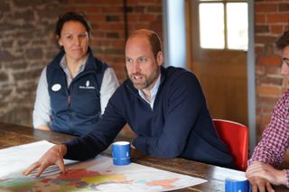 Prince William wearing a blue sweater leaning across a table and talking while touching a map with a woman in a blue vest sitting to one side and a man in a checkered shirt on the other