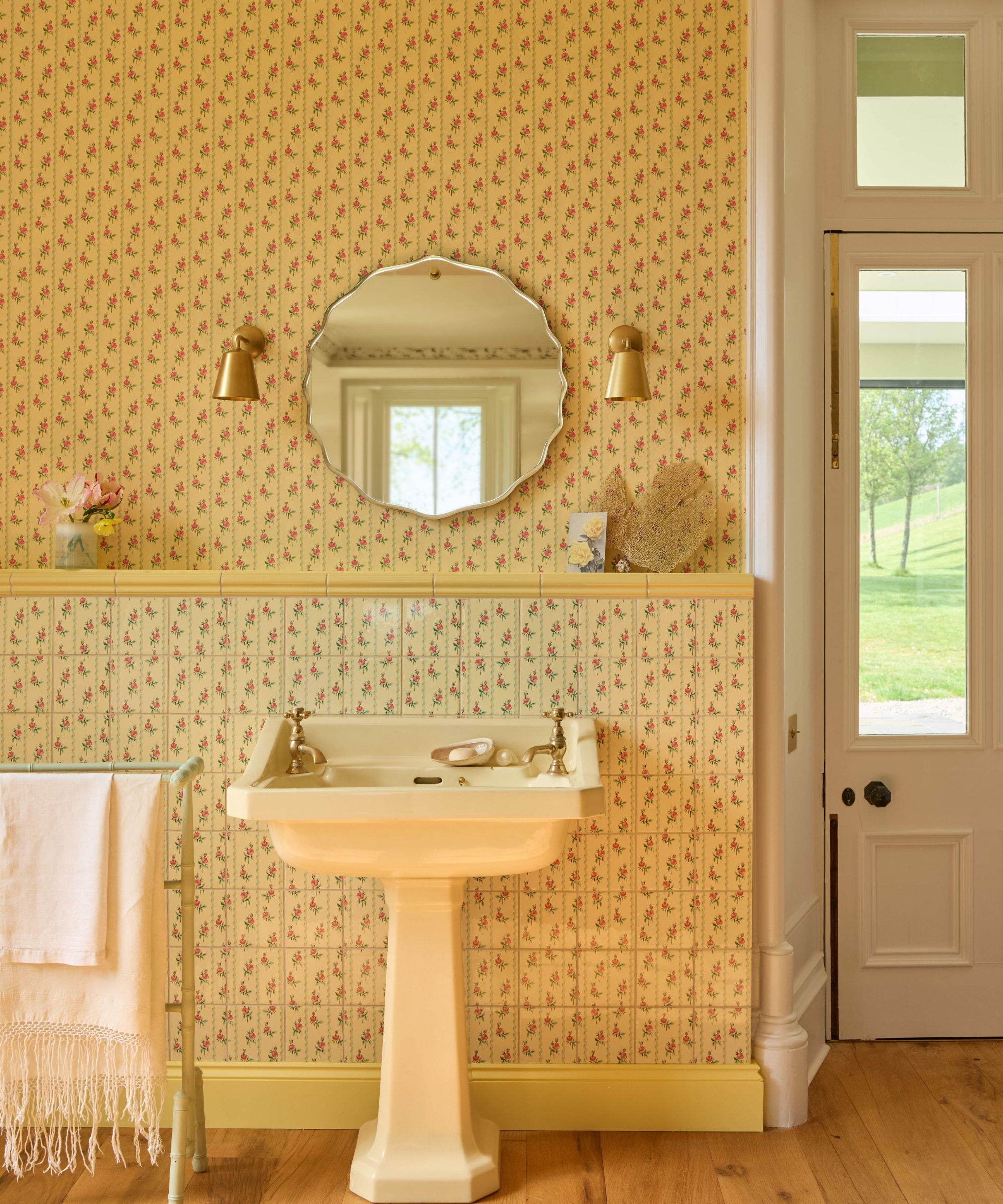 A yellow wallpapered country-style bathroom, with a pedestal sink, a circular mirror above, and two bras pendants lights either side.