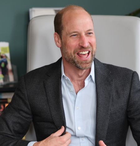 Prince William wearing a gray suit coat sitting in a chair and laughing 