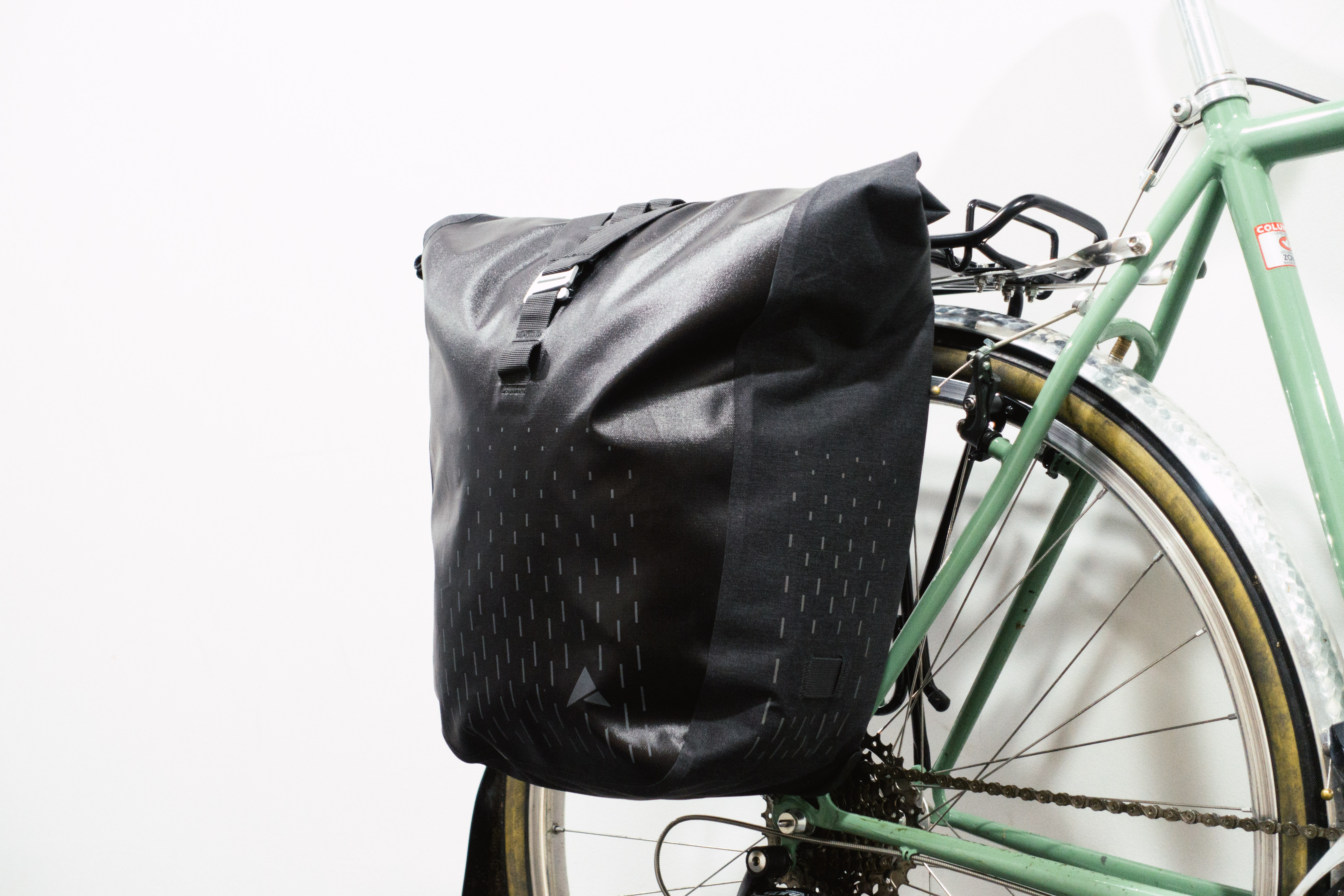 A black rolltop pannier mounted to a green bike against a white background