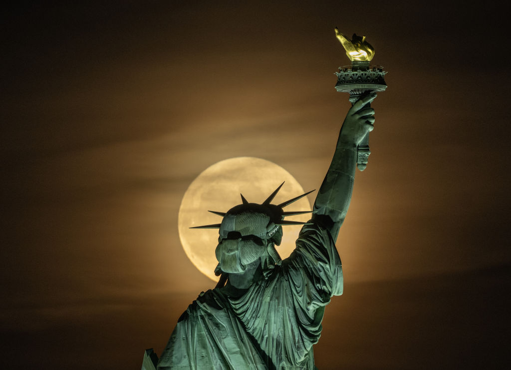 The full moon shines behind the head of the Statue of Liberty, making it look like a halo.