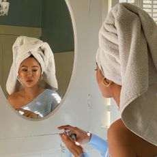 Content creator Aimee Song looking in the bathroom mirror and wearing a towel around her hair