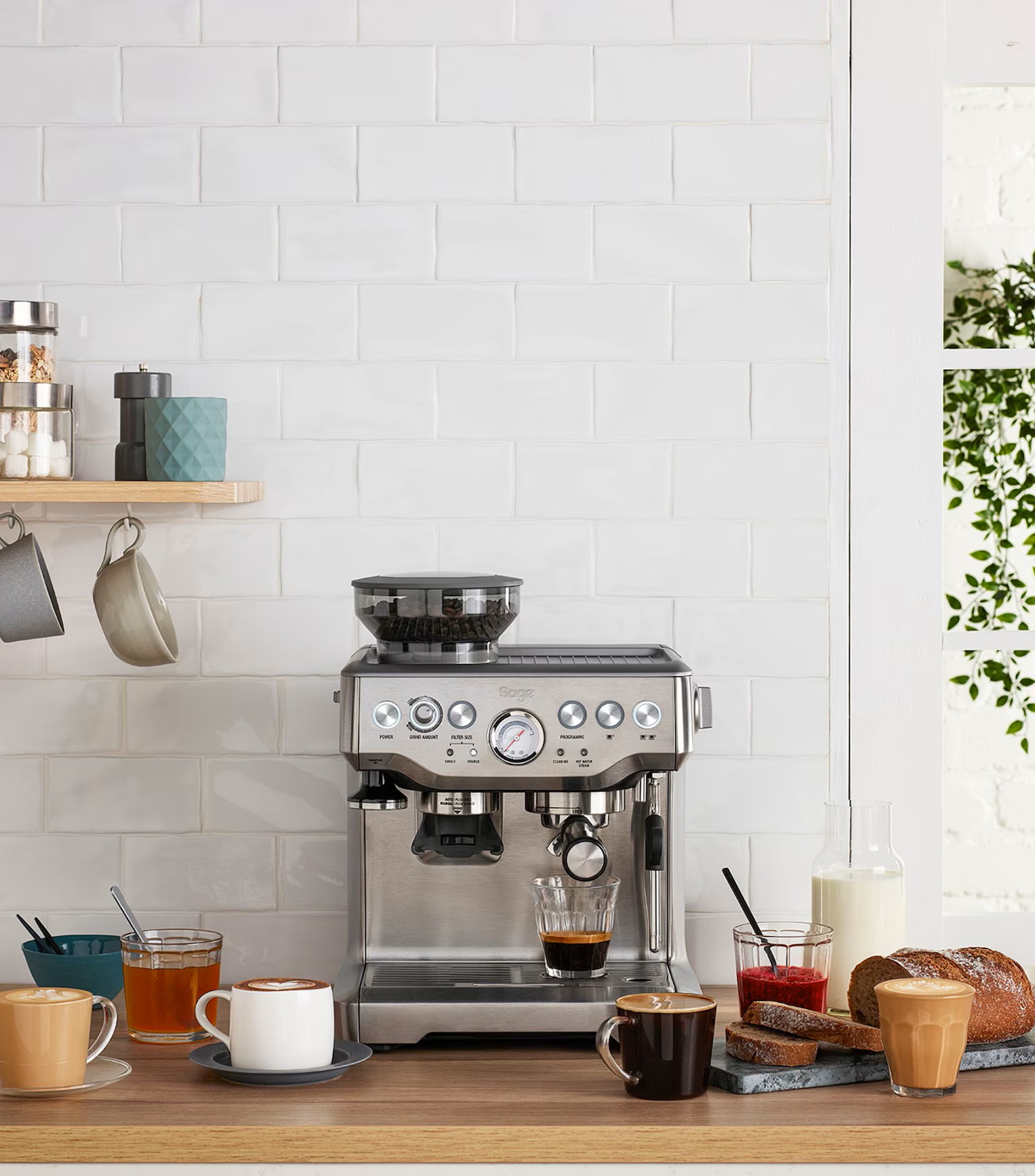 Image of a stainless steel espresso make on a wooden countertop in front of a white tile backsplash. There are coffee mugs and cups scattered around the countertop, as well as a sliced loaf of bread.