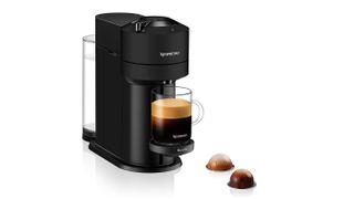 Kaffemaskinen Nespresso Vertuo Next brygger kaffe, sett mot en hvit bakgrunn med to kaplser liggende ved siden av.